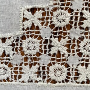 May include: Close-up of a white, intricately crocheted lace textile. The design features floral and geometric patterns, with a combination of solid and openwork sections. The lace is set against a wooden surface.