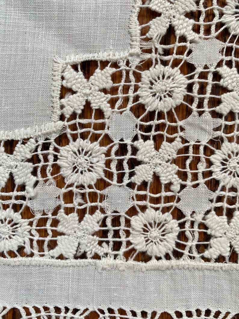 May include: Close-up of a white, intricately crocheted lace textile. The design features floral and geometric patterns, with a combination of solid and openwork sections. The lace is set against a wooden surface.