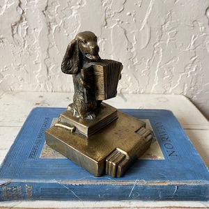 May include: A bronze-colored dog figurine playing an accordion, atop a stack of books. The figurine is detailed with visible fur and facial features. The books are on a blue book with the word "NORWAY" visible.