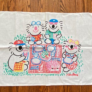 Könnte beinhalten: Ein weißes, rechteckiges Geschirrtuch mit einer farbenfrohen grafischen Illustration von vier Koalas, die ein Picknick auf einer rot karierten Decke machen. Das Design zeigt grünes Gras, Teller, Speisen und die Signatur Ken Done.