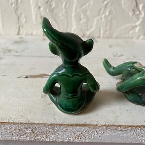 Op de afbeelding: Twee kleine, smaragdgroene keramische beeldjes. Elk beeldje heeft een zittende pose met een gebogen hoofd en puntige oren. De beeldjes staan op een wit, getextureerd oppervlak.