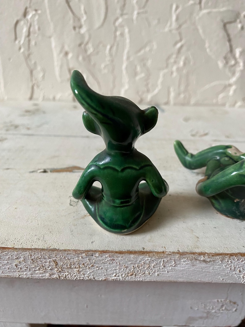 Op de afbeelding: Twee kleine, smaragdgroene keramische beeldjes. Elk beeldje heeft een zittende pose met een gebogen hoofd en puntige oren. De beeldjes staan op een wit, getextureerd oppervlak.