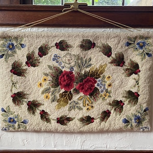 May include: A rectangular hooked rug wall hanging featuring a cream-colored background with a central floral arrangement of red roses, yellow blossoms, and blue flowers, surrounded by a leafy vine border. The text 40 and 1979 is visible.