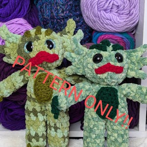 May include: Two crocheted axolotl plushies with green and brown camouflage patterns. The axolotls have red mouths, black eyes, and green feet. The plushies are sitting in a white box with yarn balls in the background. The text "PATTERN ONLY!" is superimposed on the image.