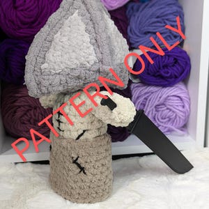 May include: A crocheted plush toy shaped like a mushroom with a gray and white hat and a brown body. The toy is holding a black knife and is surrounded by balls of yarn in various shades of purple.