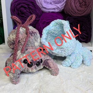 May include: Two crocheted amigurumi spider plushies, one in a light blue and white color scheme and the other in a pink and gray color scheme. The plushies are sitting on a white fluffy surface. The text "PATTERN ONLY" is superimposed on the image.