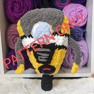 Puede incluir: Un juguete de peluche tejido a crochet con forma de robot amarillo y negro con cabeza gris y visera azul. El juguete está sentado sobre una superficie blanca y esponjosa. El texto "PATTERN ONLY" está superpuesto en la imagen.