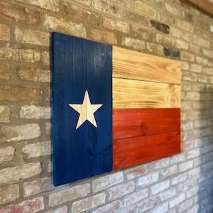 Peut inclure: Décoration murale en bois représentant le drapeau du Texas. Le drapeau présente une bande verticale bleue avec une étoile blanche et des rayures horizontales en bois naturel, jaune et rouge. L'œuvre est fixée sur un mur de briques.