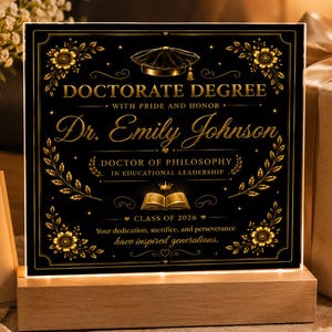 May include: A black and gold Doctorate Degree plaque with the text "Dr. Emily Johnson" and "Class of 2026". The plaque is illuminated by a wooden base. A card reads "Congratulations Doctor! We are so proud of you!"