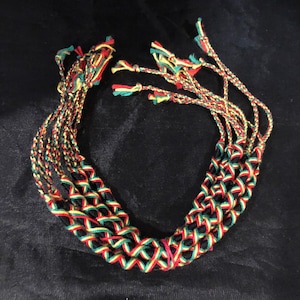 May include: A set of eight friendship bracelets in black, red, yellow, and green colors. The bracelets are woven together in a pattern.