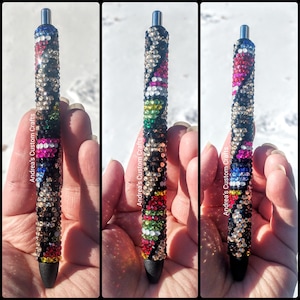 May include: Three black pens with a silver top and a black tip. Each pen is covered in colorful rhinestones in a geometric pattern. The pens are held in a hand against a white background.