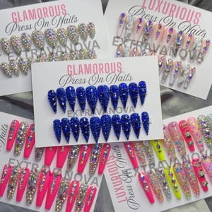 May include: Five sets of press-on nails displayed on white cards labeled Glamorous Press On Nails or Luxurious Press On Nails with the brand name EXTRAVANOVA. The sets feature various designs including blue, pink, and nude bases with rhinestones.