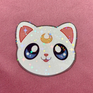 May include: A white cat sticker with a crescent moon on its forehead and sparkly blue eyes. The sticker has a holographic finish and is covered in glitter.