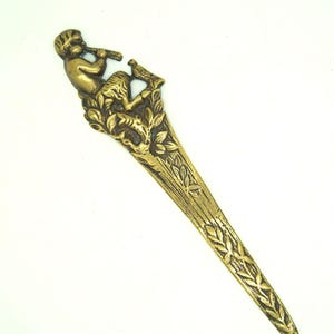 May include: A gold-toned metal letter opener featuring an intricate design of a figure playing a flute and a bird perched on a branch. The handle is shaped like a bird and the blade is long and slender.
