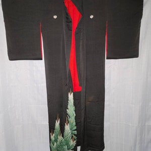Pode incluir: Um quimono de seda preto com forro vermelho e bordado floral verde e dourado. O quimono tem duas flores brancas bordadas no peito.