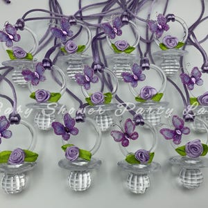 May include: Clear pacifiers with purple butterfly and rose decorations. Each pacifier has a clear ring and a decorative purple cord. The text "Baby Shower Party Land" is visible in the image.