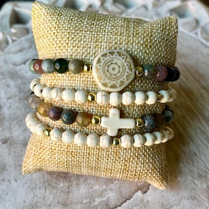 May include: A set of four bracelets featuring white beads, gold accents, and a cross charm. The bracelets are stacked on a beige fabric surface.