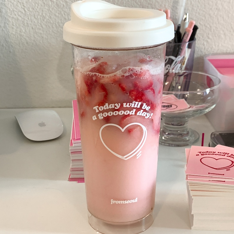 以下が含まれることがあります： 白い蓋が付いた透明なガラスのタンブラーに、イチゴが入ったピンク色の飲み物が入っています。タンブラーには「Today will be a gooooood day!」と「fromseoul」の文字が印刷されています。