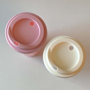 以下が含まれることがあります： ふた付きの使い捨てコーヒーカップが2つ。左側のカップはピンクで、右側のカップは白です。