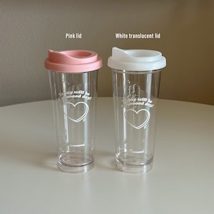 以下が含まれることがあります： ふた付きの透明なプラスチック製のタンブラーが2つ。左側のタンブラーはピンクのふた、右側のタンブラーは白いふた。両方のタンブラーには「Today will be a good day!」という文字とハートのデザインが側面に印刷されています。「fromseoul」という文字が各タンブラーの底に印刷されています。