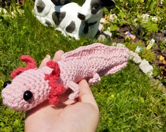 May include: A light pink crocheted axolotl plush toy featuring a long body, a flat tail, and small limbs. The creature has two prominent black plastic eyes and bright red, textured external gills extending from the sides of its head.