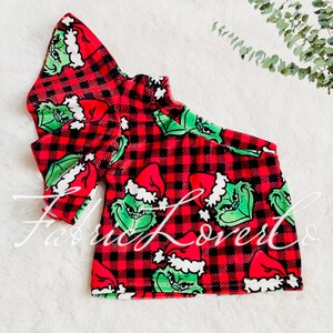 May include: Red and black plaid one-shoulder top with a green Grinch print. The top has a ruffle on one sleeve.