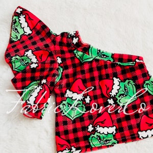 May include: Red and black plaid fabric with a green Grinch character wearing a Santa hat. The fabric is designed for clothing and features a repeating pattern.