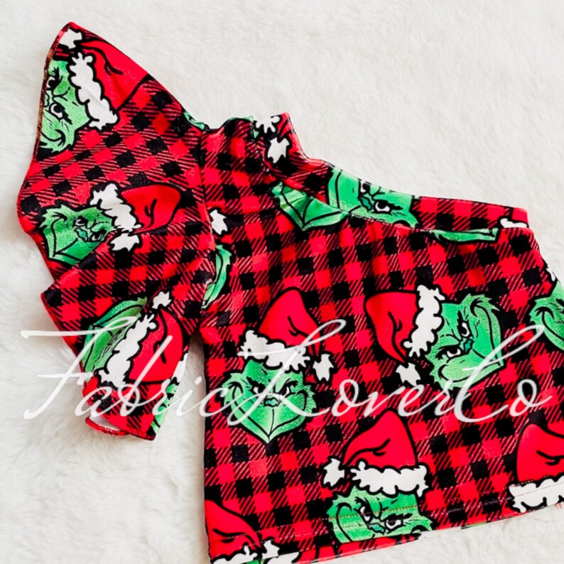 May include: Red and black plaid fabric with a green Grinch character wearing a Santa hat. The fabric is designed for clothing and features a repeating pattern.