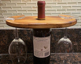 May include: A wooden wine glass holder with a bottle of Rutherford Cabernet Sauvignon. The holder has a circular top with two cutouts for wine glasses and a hole for the bottle. Two wine glasses are suspended below the wooden top.