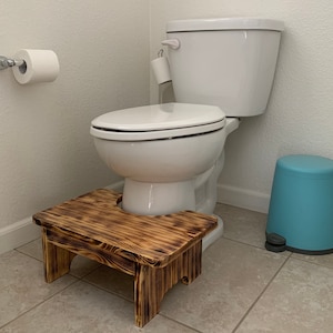 May include: A wooden step stool with a burnt wood finish sits in front of a white toilet. The step stool has a rectangular top and four legs. A blue trash can is to the right of the toilet. A roll of toilet paper is on the wall.