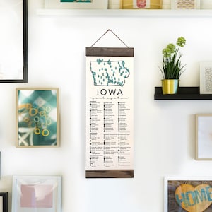 May include: A white wall gallery with a variety of framed artwork, including a map of Iowa with a list of cities, a butterfly, a heart with the word "home", and abstract designs. The gallery is decorated with plants and shelves.