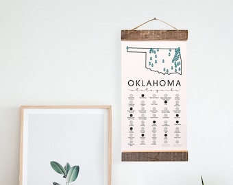 May include: A white wall with a wooden framed print of a map of Oklahoma with the text "OKLAHOMA state parks" and a list of state parks. The print is hanging on the wall above a white dresser with two drawers. A white vase with a green plant is on the dresser. A white moon lamp is on the dresser to the right of the vase.
