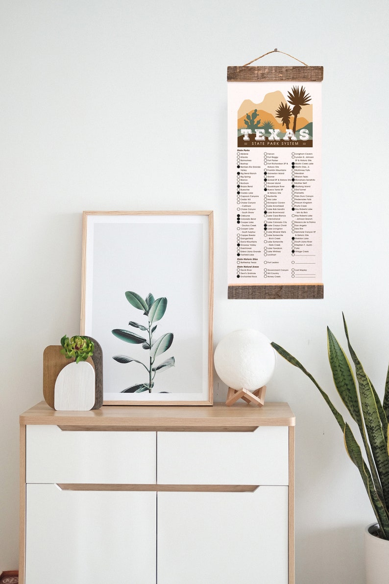 May include: A printable checklist of Texas State Parks. The checklist is printed on a light pink background with brown text and black checkboxes. The checklist is mounted on a wooden dowel rod with a brown rope hanger. The checklist is hanging on a white wall with a framed print of a green plant and a white moon lamp on a wooden stand.