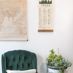 May include: A checklist of Texas State Parks, printed on a canvas scroll, hanging on a white wall. The checklist includes the name of each park and a box to check off once visited. The scroll is hanging above a green velvet armchair with a striped pillow.