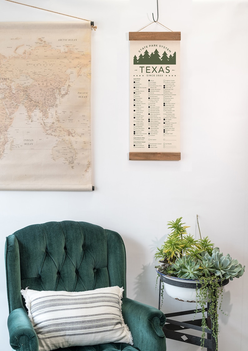 May include: A checklist of Texas State Parks, printed on a canvas scroll, hanging on a white wall. The checklist includes the name of each park and a box to check off once visited. The scroll is hanging above a green velvet armchair with a striped pillow.