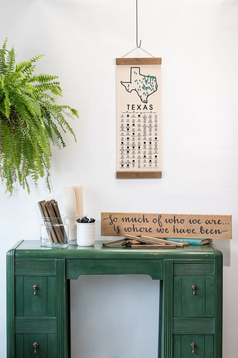 May include: A green desk with drawers and a wooden sign that reads "So much of who we are... Is where we have been." A canvas print of the state of Texas with the word "Texas" printed below the map hangs on the wall above the desk.