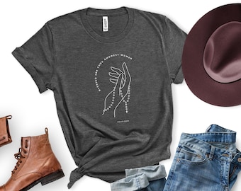 Puede incluir: Una camiseta gris oscuro con un gráfico blanco de manos sosteniendo un rosario y el texto "Blessed are thou amongst women".