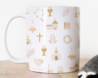 May include: White ceramic mug with a pattern of gold and pink religious symbols, including a cross, a church, a chalice, and a rosary. The mug is on a wooden surface.