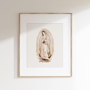 Op de afbeelding: Een ingelijste print van een aquarel die Onze Lieve Vrouw van Guadalupe afbeeldt, een religieuze figuur in een blauwe en witte jurk, staand op een halve maan.