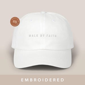Op de afbeelding: Witte baseballpet met de tekst "WALK BY FAITH" in wit geborduurd op de voorkant.