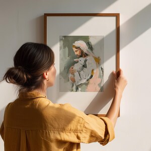 Peut inclure: Une impression d'art encadr&eacute;e repr&eacute;sentant une figure religieuse tenant un agneau, avec des coups de pinceau doux dans des tons de blanc, rose et vert. L'&oelig;uvre est tenue par une personne portant une chemise jaune, contre un mur blanc.