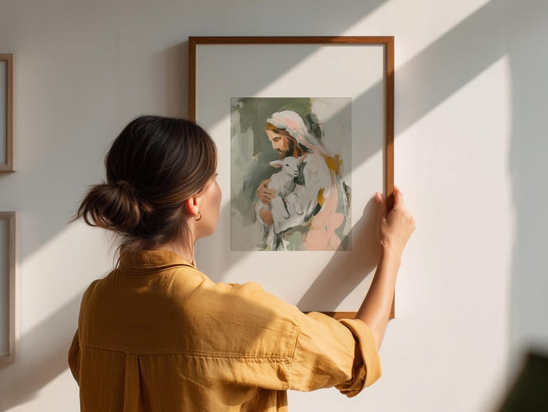 Peut inclure: Une impression d'art encadr&eacute;e repr&eacute;sentant une figure religieuse tenant un agneau, avec des coups de pinceau doux dans des tons de blanc, rose et vert. L'&oelig;uvre est tenue par une personne portant une chemise jaune, contre un mur blanc.