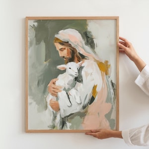 Peut inclure: Une impression d'art encadr&eacute;e repr&eacute;sentant une figure en robe blanche tenant un agneau. La peinture utilise une palette douce de verts, de roses et de jaunes. L'&oelig;uvre est tenue par une personne portant une chemise blanche.