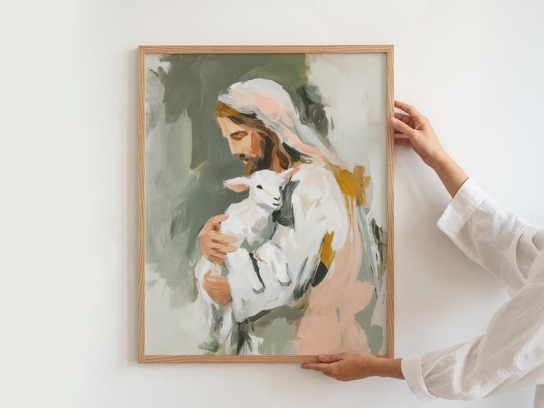 Peut inclure: Une impression d'art encadr&eacute;e repr&eacute;sentant une figure en robe blanche tenant un agneau. La peinture utilise une palette douce de verts, de roses et de jaunes. L'&oelig;uvre est tenue par une personne portant une chemise blanche.