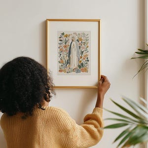 May include: A framed art print featuring a watercolor illustration of a religious figure surrounded by flowers. The artwork is in a gold frame and is being held up against a white wall. The color palette includes soft blues, greens, and yellows.