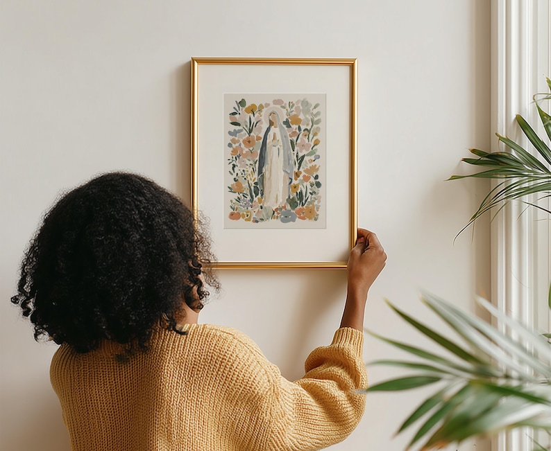 May include: A framed art print featuring a watercolor illustration of a religious figure surrounded by flowers. The artwork is in a gold frame and is being held up against a white wall. The color palette includes soft blues, greens, and yellows.