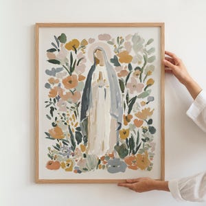 May include: A framed art print featuring a religious figure surrounded by colorful flowers. The artwork depicts a woman in a white and blue robe with a halo, set against a backdrop of peach, yellow, and green floral elements. The frame is made of light-colored wood.