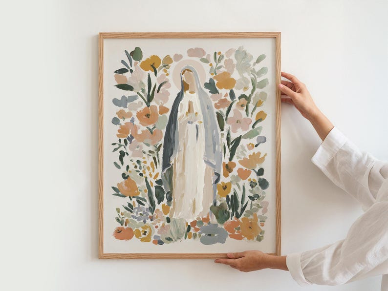 May include: A framed art print featuring a religious figure surrounded by colorful flowers. The artwork depicts a woman in a white and blue robe with a halo, set against a backdrop of peach, yellow, and green floral elements. The frame is made of light-colored wood.