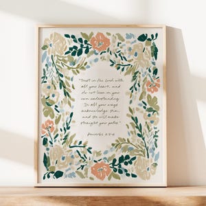 May include: A framed art print with a floral wreath design in peach, green, and blue hues. The print displays the text "Trust in the Lord with all your heart..." from Proverbs 3:5-6. The frame is a light wood colour.