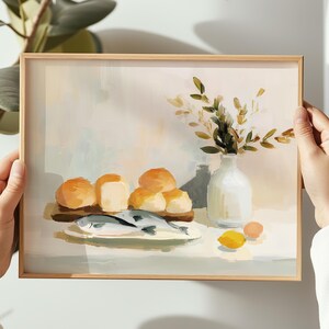 May include: A framed art print depicting a still life scene. The painting features a plate with fish, bread rolls, a vase with greenery, and a lemon. The color palette includes beige, white, and shades of orange and green.
